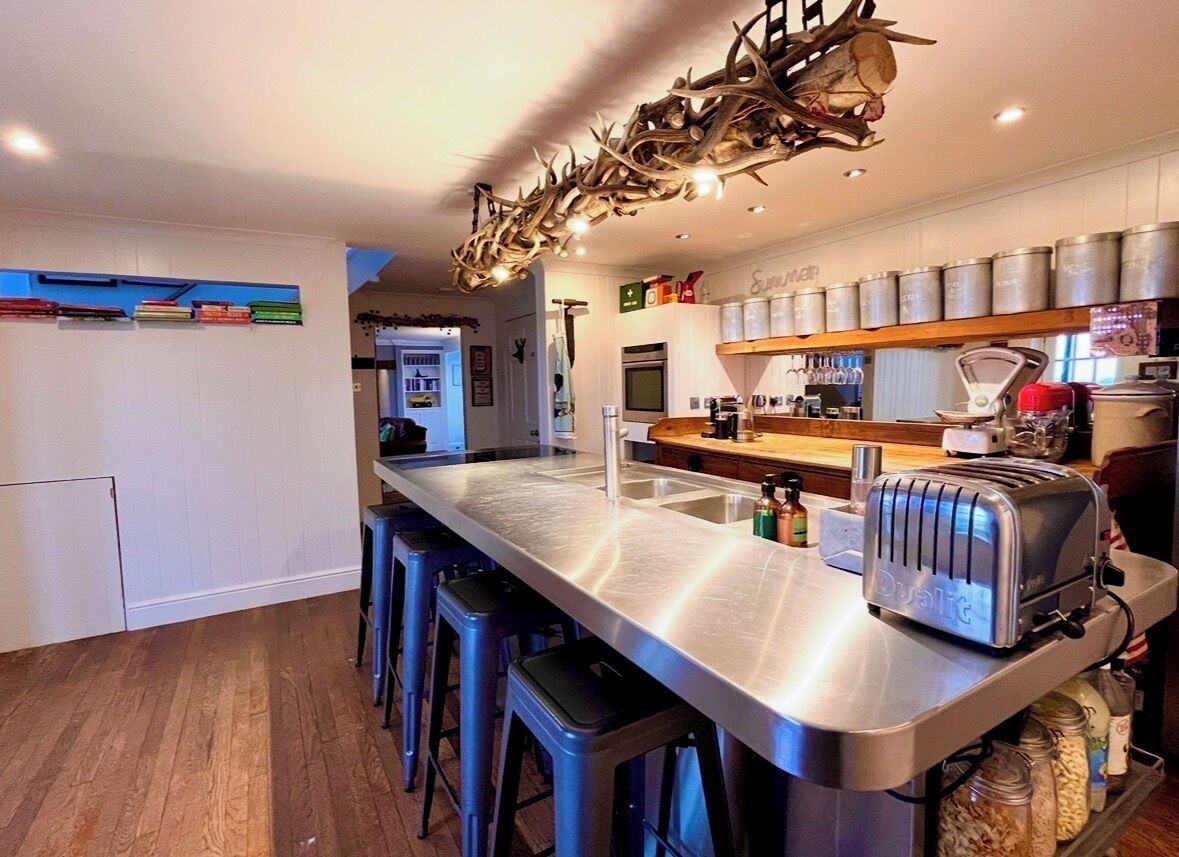 A modern kitchen with a stainless steel island, bar stools, a toaster, and a unique antler chandelier.