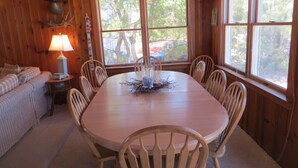 Uma sala de jantar de madeira com uma mesa redonda, seis cadeiras, um abajur e vista para árvores através de grandes janelas.