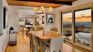 A dining area with a wooden table and chairs, a chandelier, and a large window with a view of the sea.