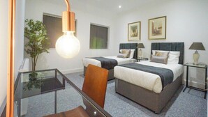 Une chambre d’hôtel avec deux lits, une table en verre, une chaise, un bureau, une lampe, une plante et des tableaux encadrés au mur.