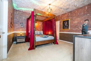 Una cama con dosel y cortinas rojas, una mesita de noche, una lámpara de pared y un cuadro enmarcado.