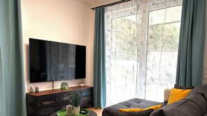 Un salon équipé d’un téléviseur à écran plat, d’un canapé avec des coussins, d’une table basse et d’une fenêtre avec des rideaux.