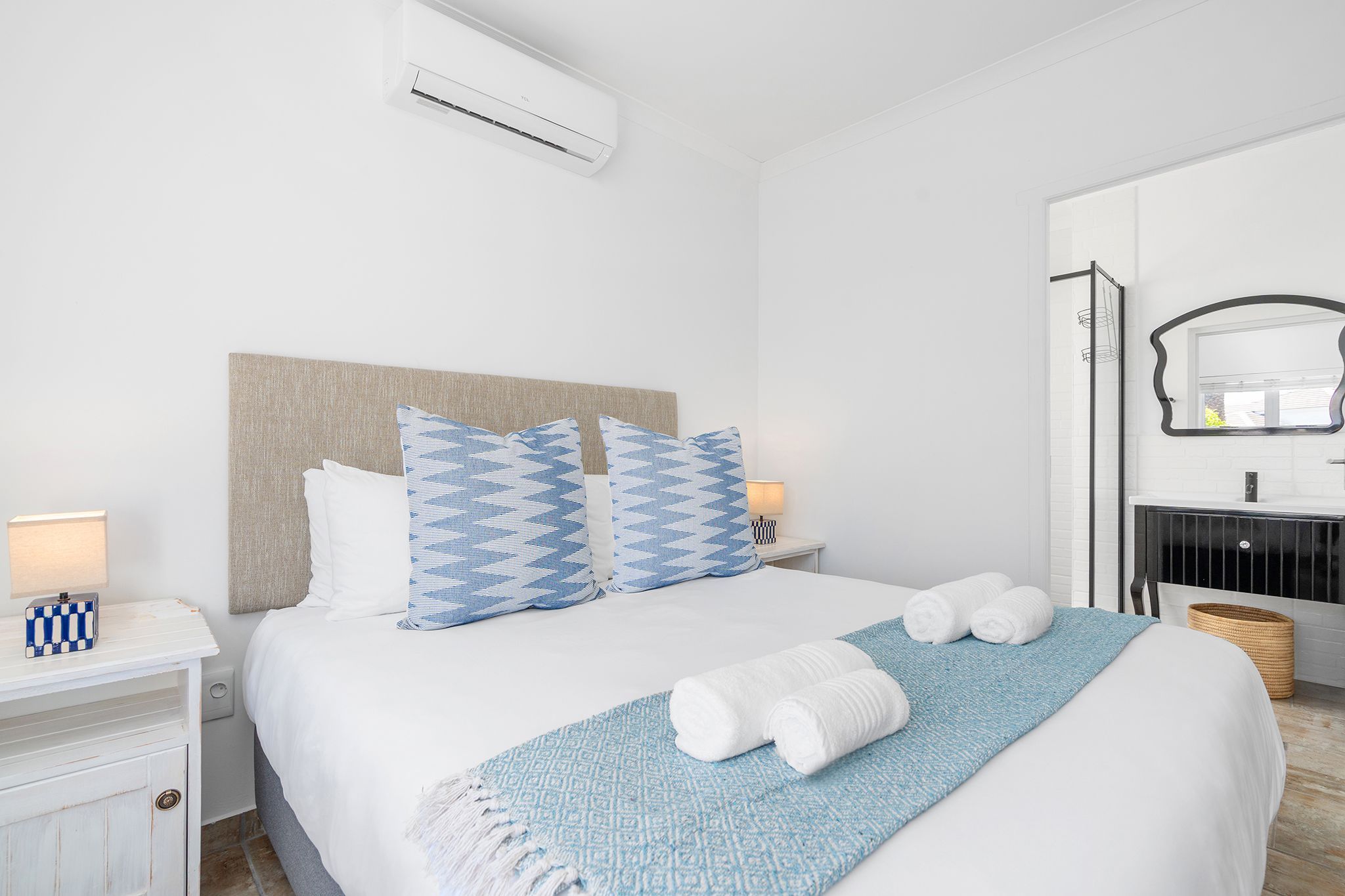A hotel room with a bed, a nightstand, a lamp, a mirror, and a bathroom visible through an open door.