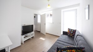 A modern living room with a grey sofa, a flat-screen TV on a white stand, a small table, and a window with blinds.