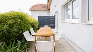 A patio with a wooden table and white chairs, surrounded by greenery and a white building with a tiled roof.
