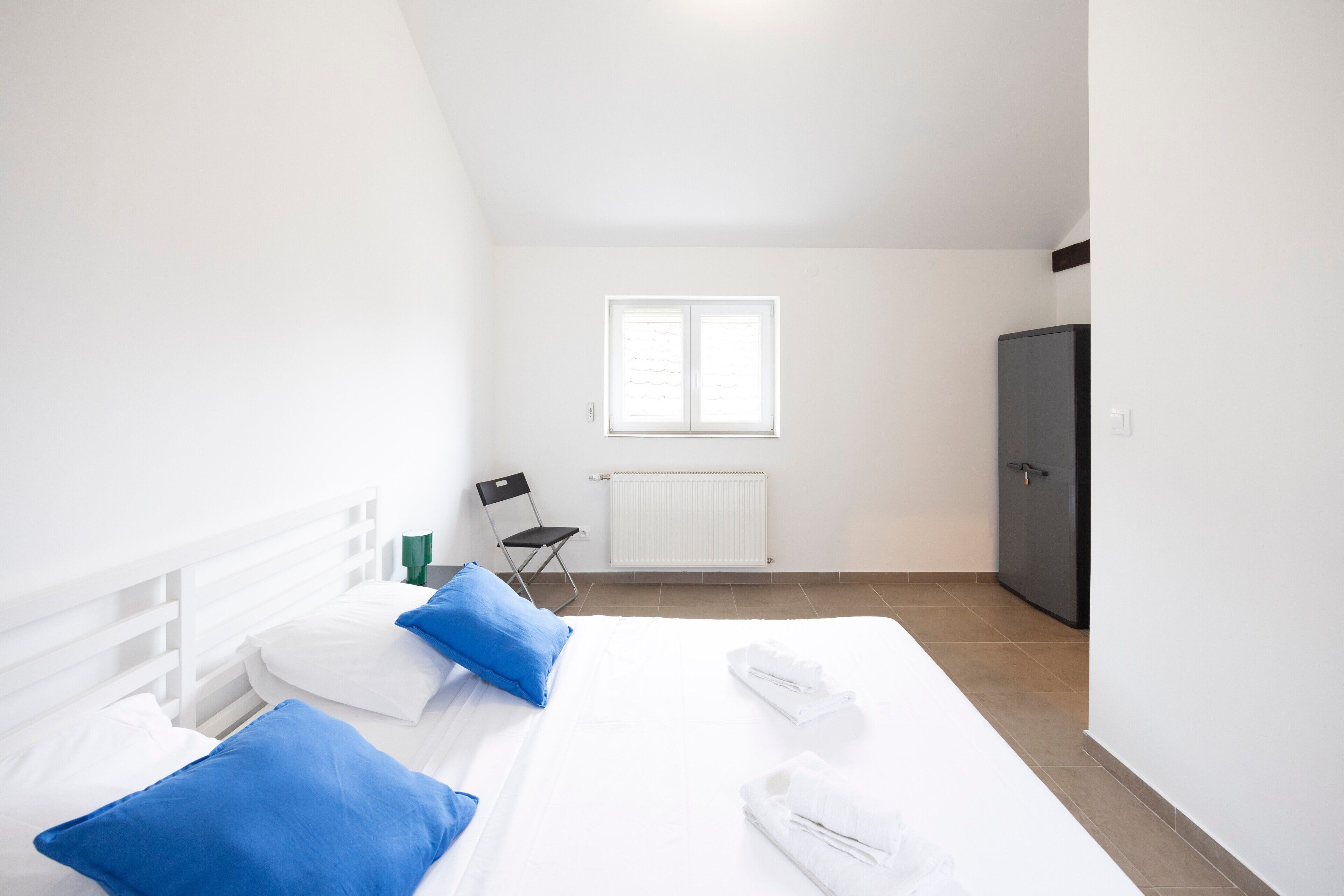 A hotel room with a bed, pillows, a radiator, a window, a chair, and a wardrobe.