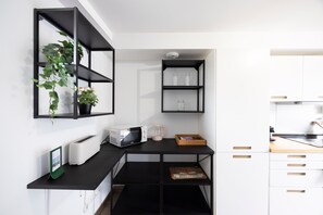 A kitchenette with a microwave, toaster, and coffee maker on a black countertop, with shelves above holding glass jars and a potted plant.