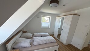 Een slaapkamer met een schuine kap, een bed met gestreept beddengoed, een houten nachtkastje, een kledingkast en een raam met uitzicht op groen.