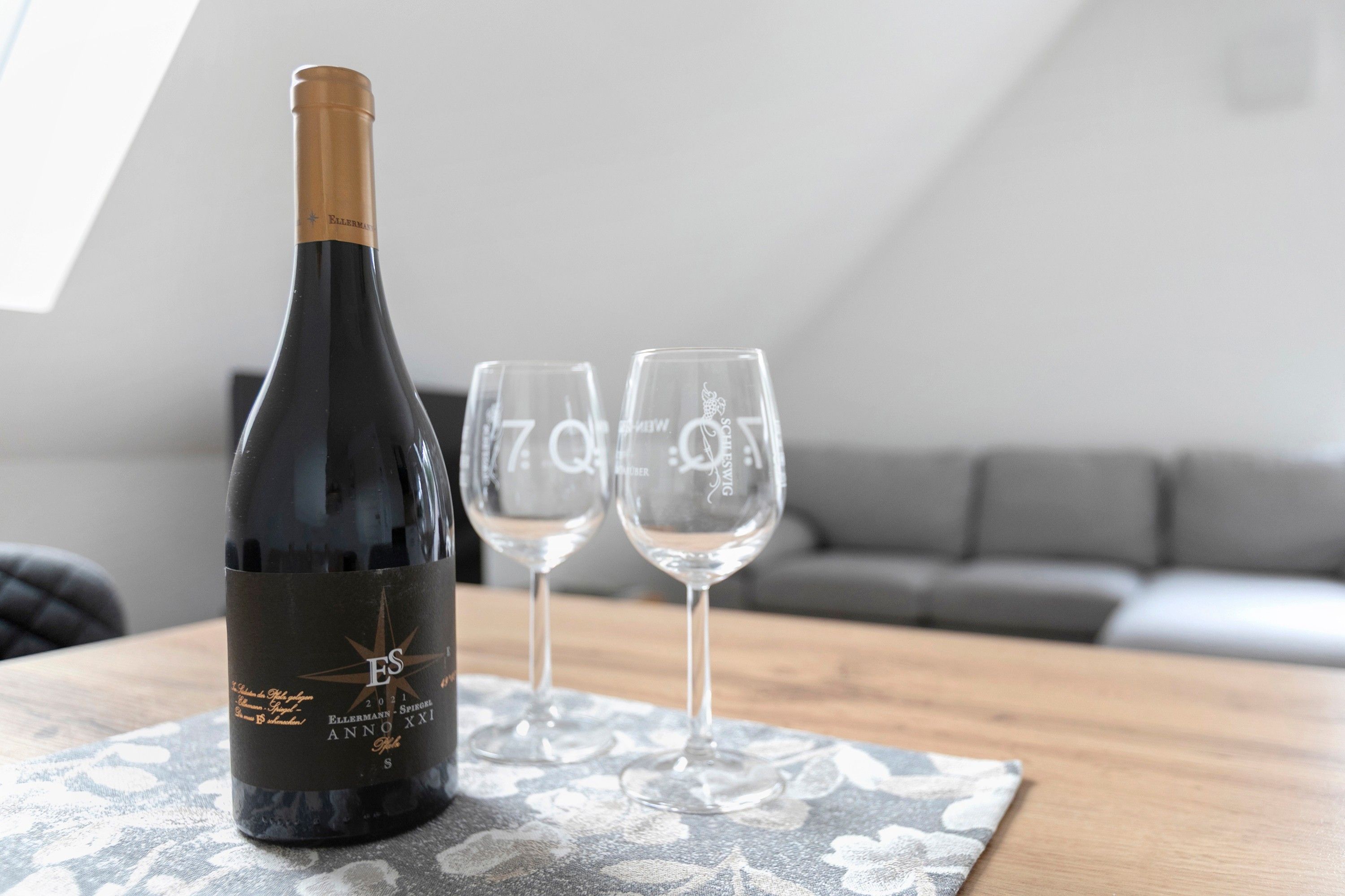 Uma garrafa de vinho e duas taças sobre uma toalha de mesa estampada em um quarto com um sofá cinzento.