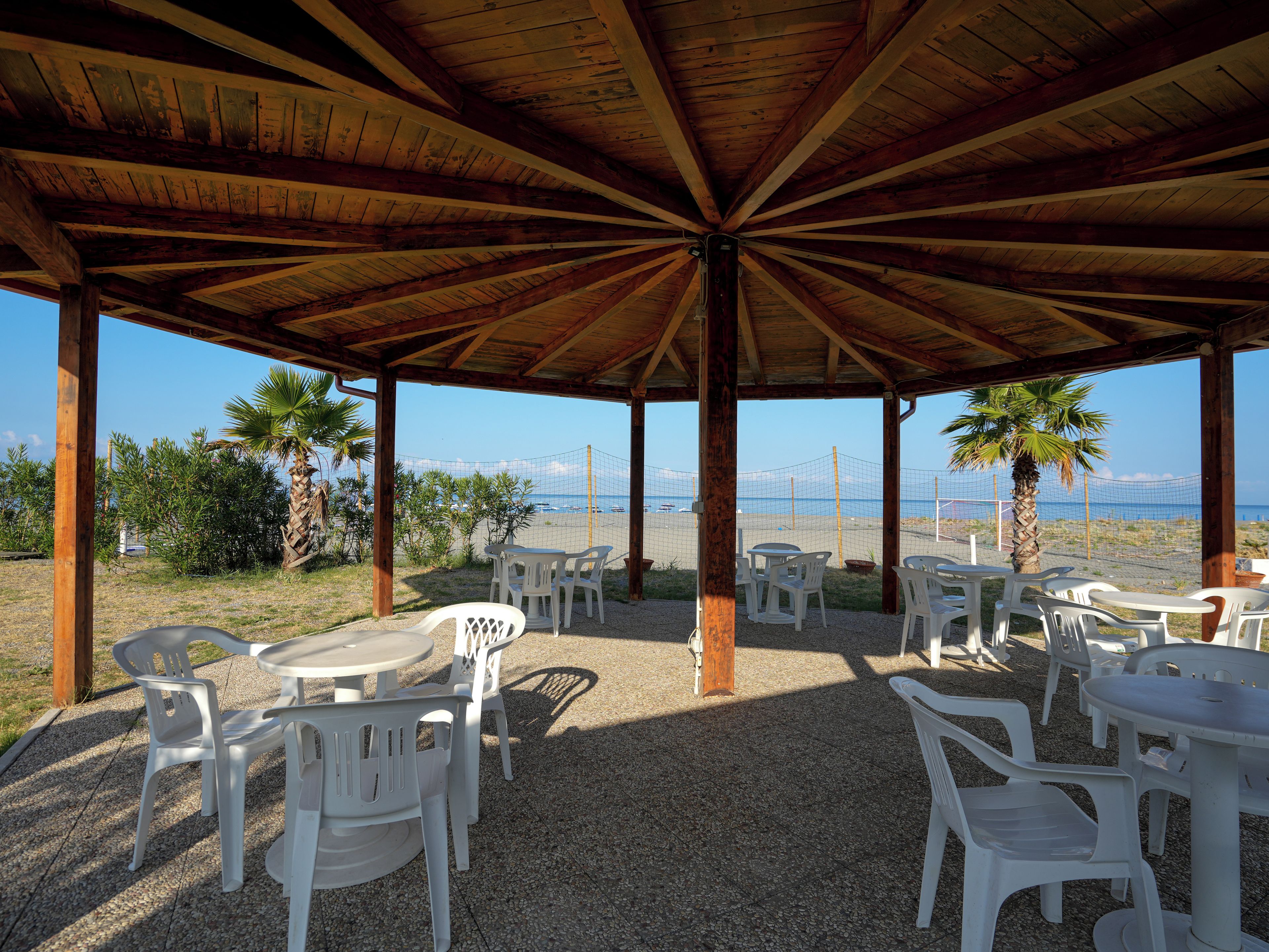 An outdoor covered seating area with tables and chairs, overlooking a beach with a volleyball net.