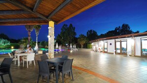 An outdoor patio area with tables and chairs, a stone pillar, and a pool in the background. - FRUIT VILLAGE HOLIDAY BEACH (Diamante)
