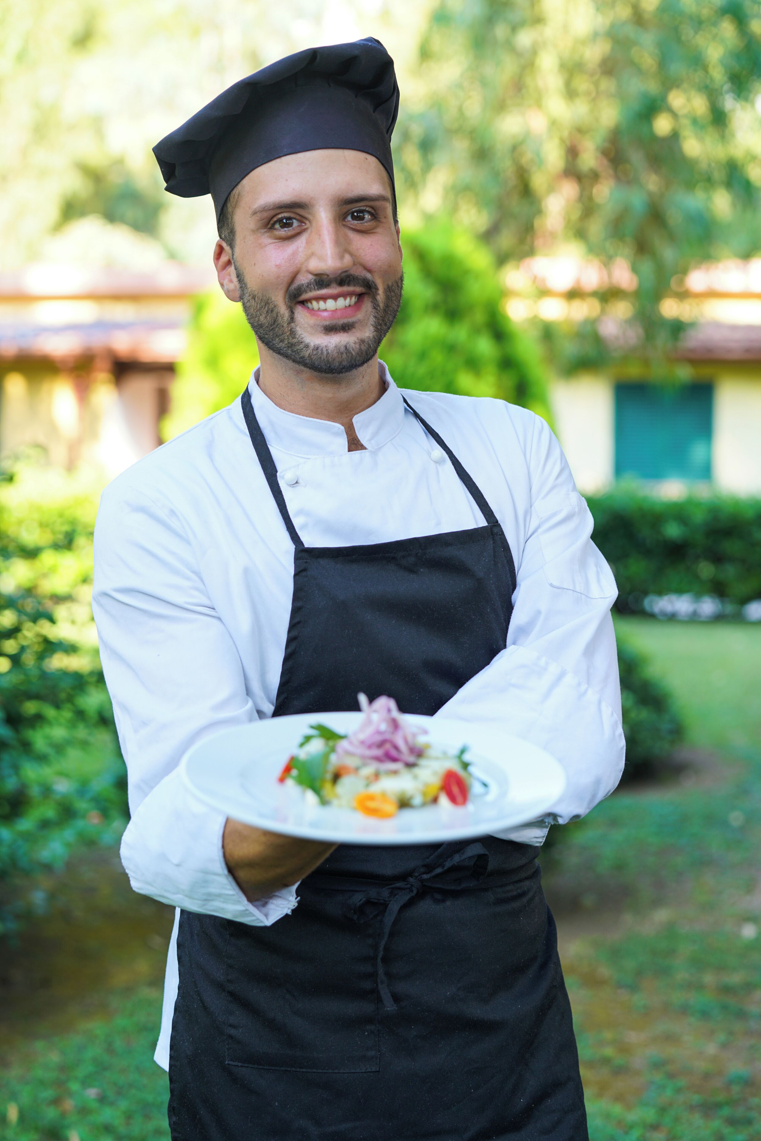 Un chef con uniforme blanco y delantal negro sostiene un plato de comida al aire libre.