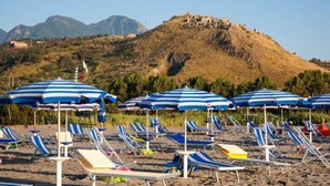 A beach with sun loungers and striped umbrellas, a hill with ruins in the background. - FRUIT VILLAGE HOLIDAY BEACH (Diamante)