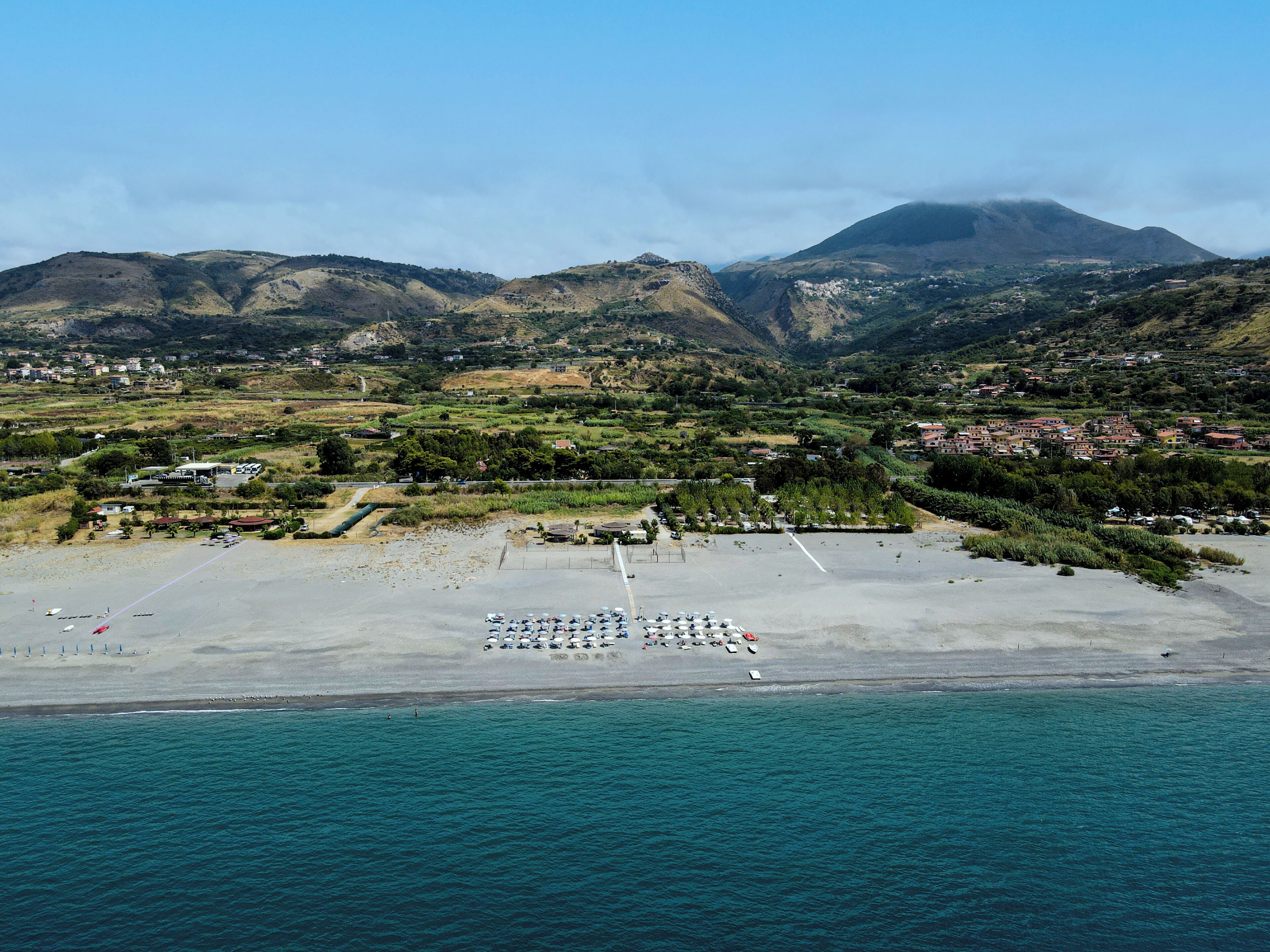 Vue aérienne d’une zone côtière avec une plage, un parking et des bâtiments en arrière-plan.