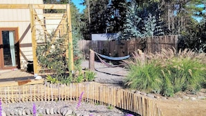 A wooden cabin with a glass door, surrounded by a garden with various plants and a hammock.