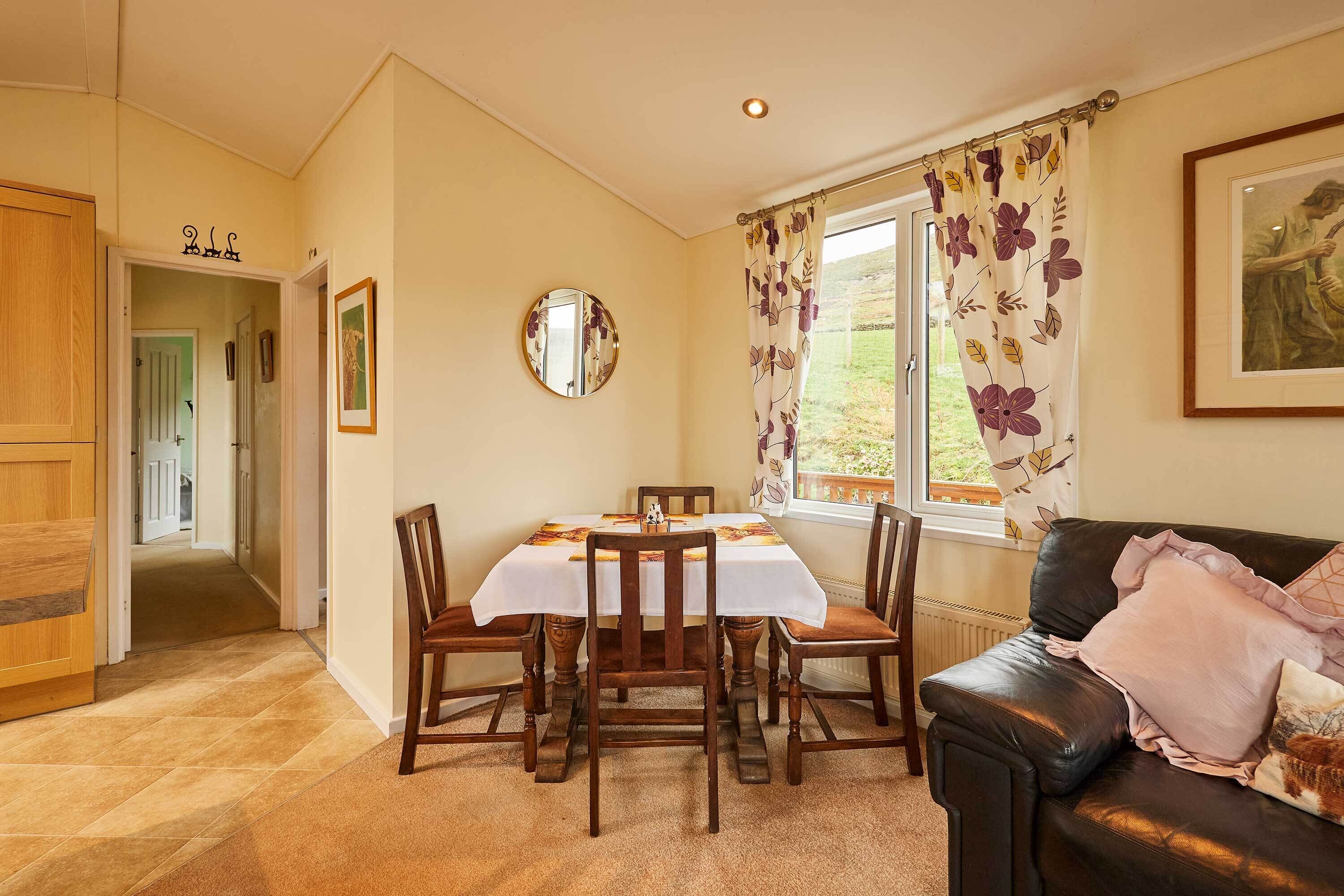 Un coin repas avec une table dressée pour six personnes, entouré de chaises en bois, d’un canapé en cuir noir et d’une fenêtre ornée de rideaux à motifs floraux.