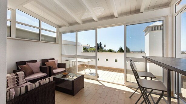 Uma sala de estar moderna com sofá, mesa de centro e área de jantar com mesa e cadeiras. Grandes janelas oferecem vista para o exterior.