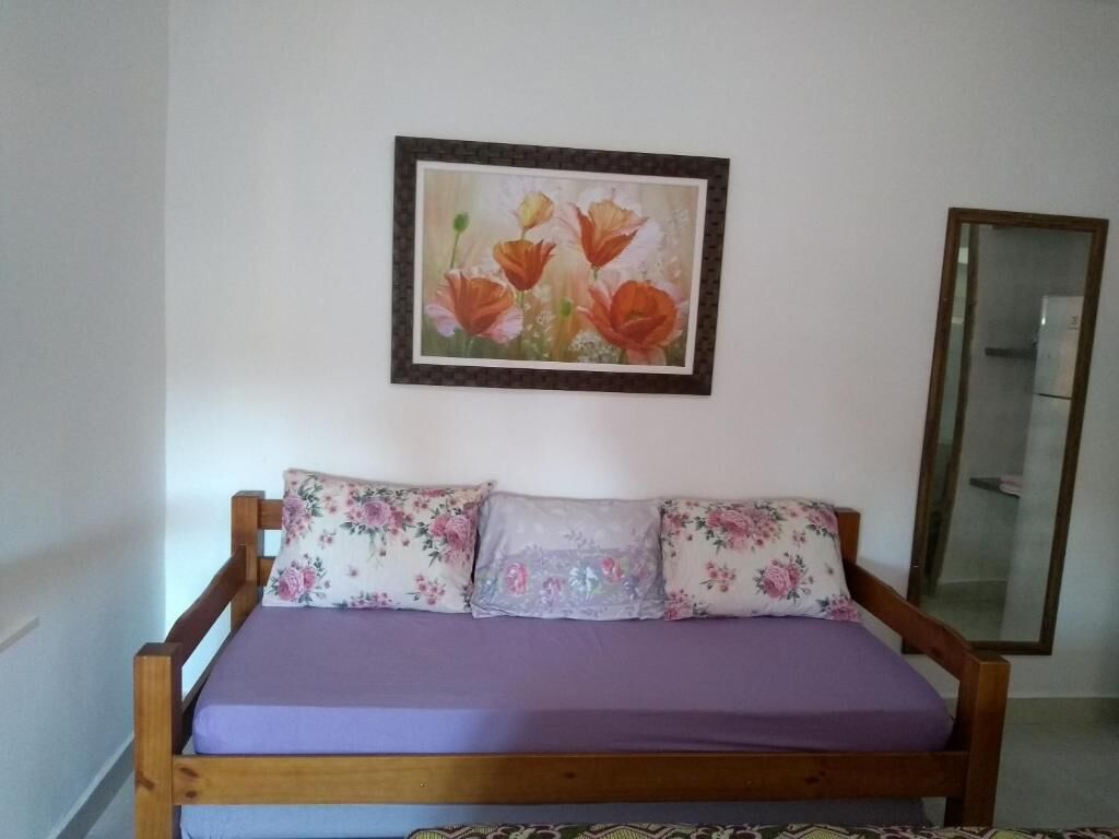 A bed with floral pillows, a purple bedspread, and a framed floral painting on the wall.