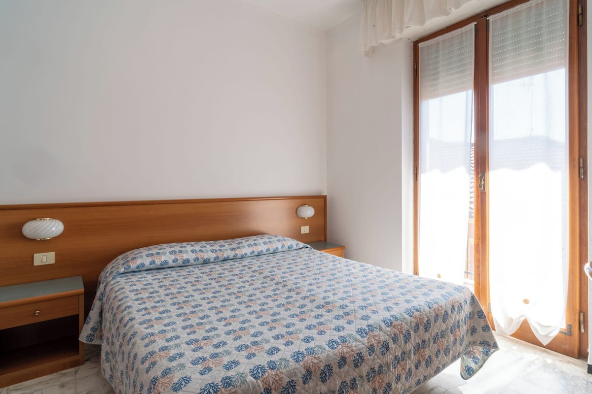 Un dormitorio con cama, mesita de noche, enchufe y ventana con cortinas.