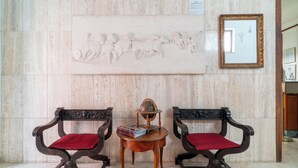 Una hall con pareti in marmo, due poltrone in legno decorate con cuscini rossi, un tavolino in legno con un mappamondo e libri, e uno specchio incorniciato appeso al muro.