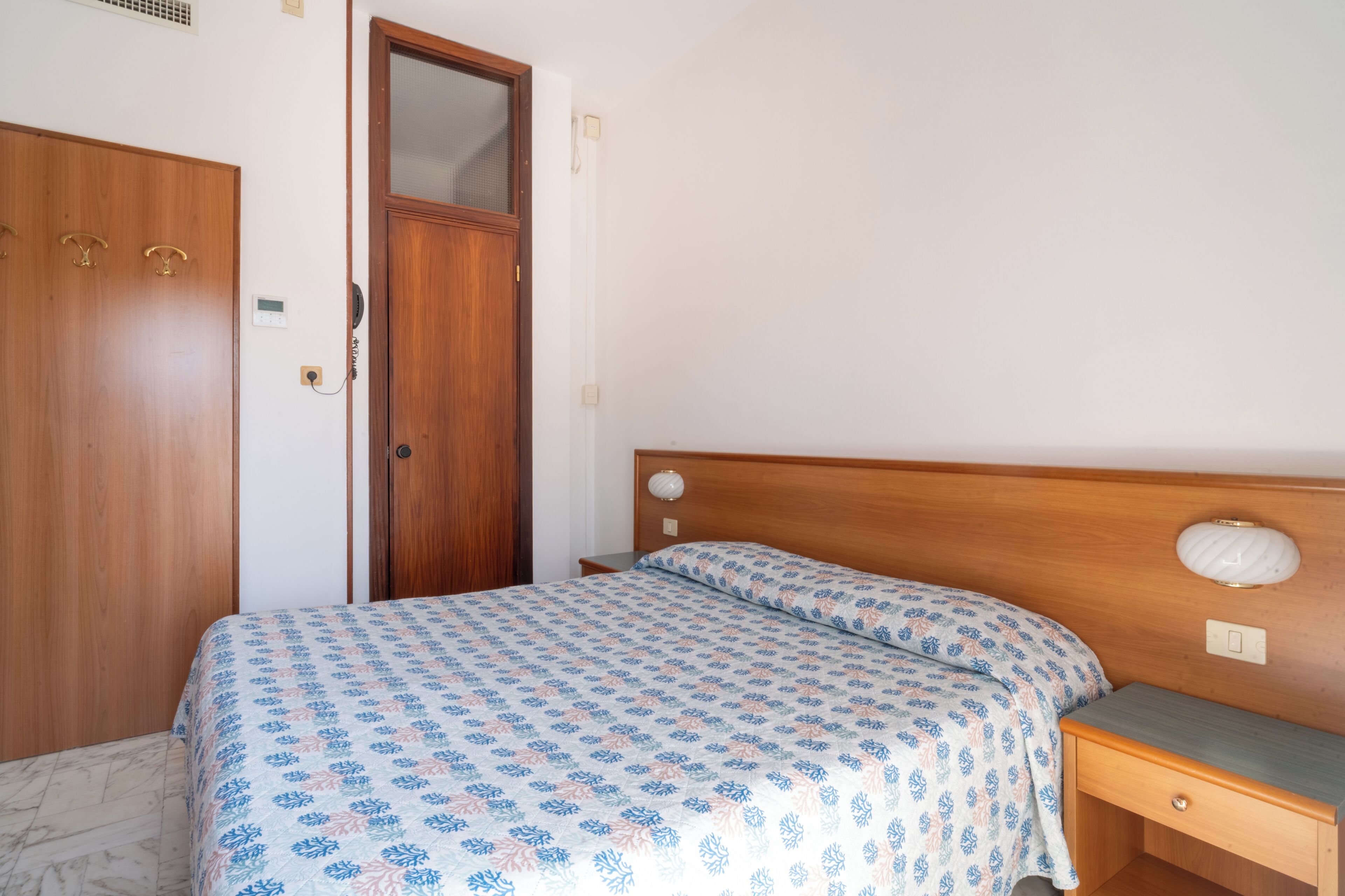 A bedroom with a wooden wardrobe, a bed with patterned bedding, a bedside table, and a nightstand.
