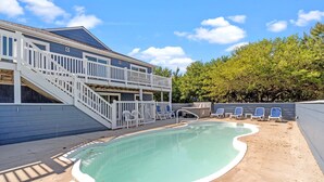 Piscina com um deck branco, área de estar coberta e escadaria que leva a um andar superior.
