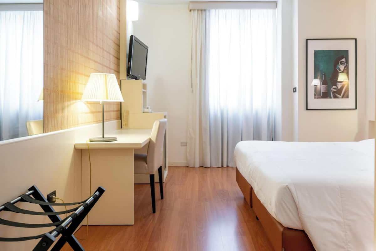 A hotel room with a bed, a desk with a chair, a television, and a painting on the wall.