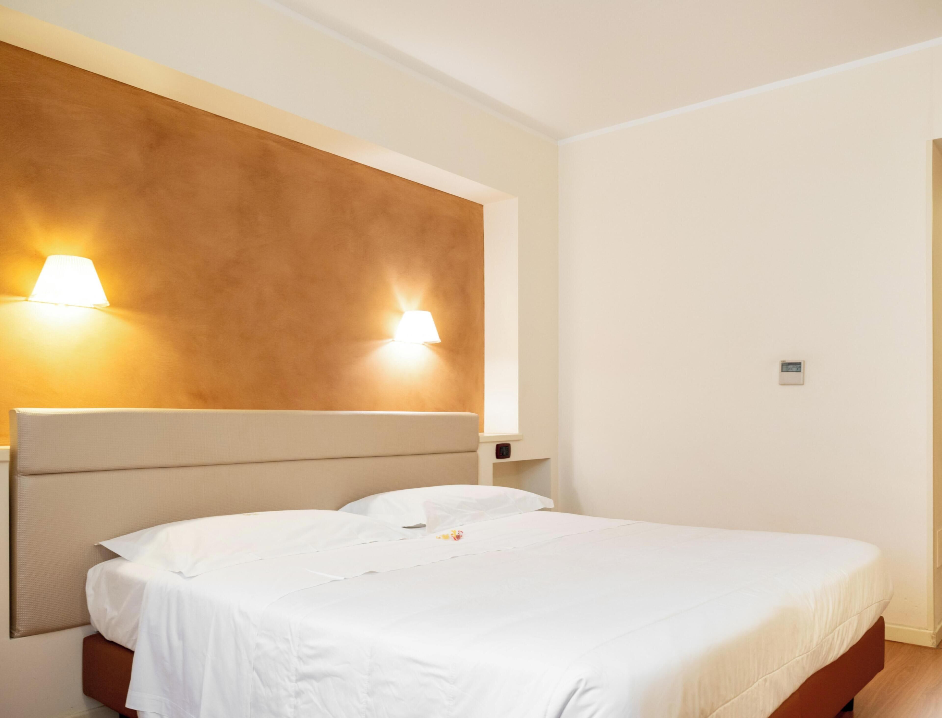 A hotel room with a bed, two wall-mounted lamps, a thermostat, and a window.
