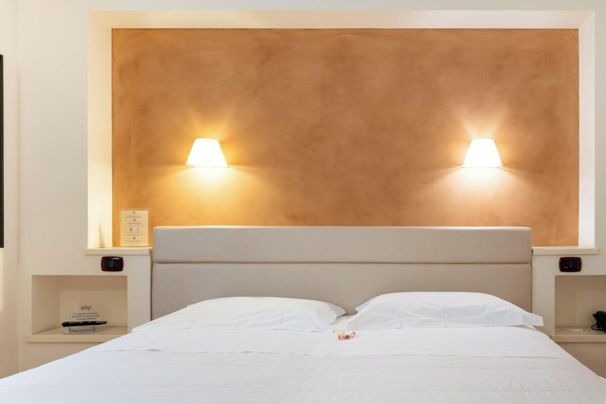 Une chambre d’hôtel avec un grand lit, deux tables de chevet, une tête de lit fixée au mur et deux lampes fixées au mur.