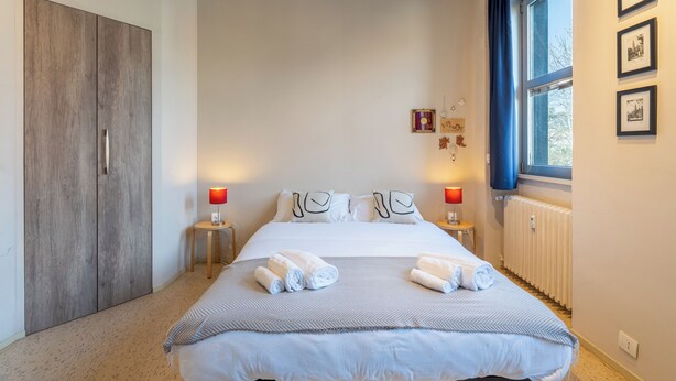 Ein Schlafzimmer mit einem Bett, zwei Nachttischen, einem Kleiderschrank, einem Fenster mit Vorhängen und Wandbildern.