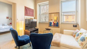Ein modernes Wohnzimmer mit einem Flachbildfernseher, einer Couch, einem blauen Sessel und einem großen Fenster mit Blick auf Gebäude.