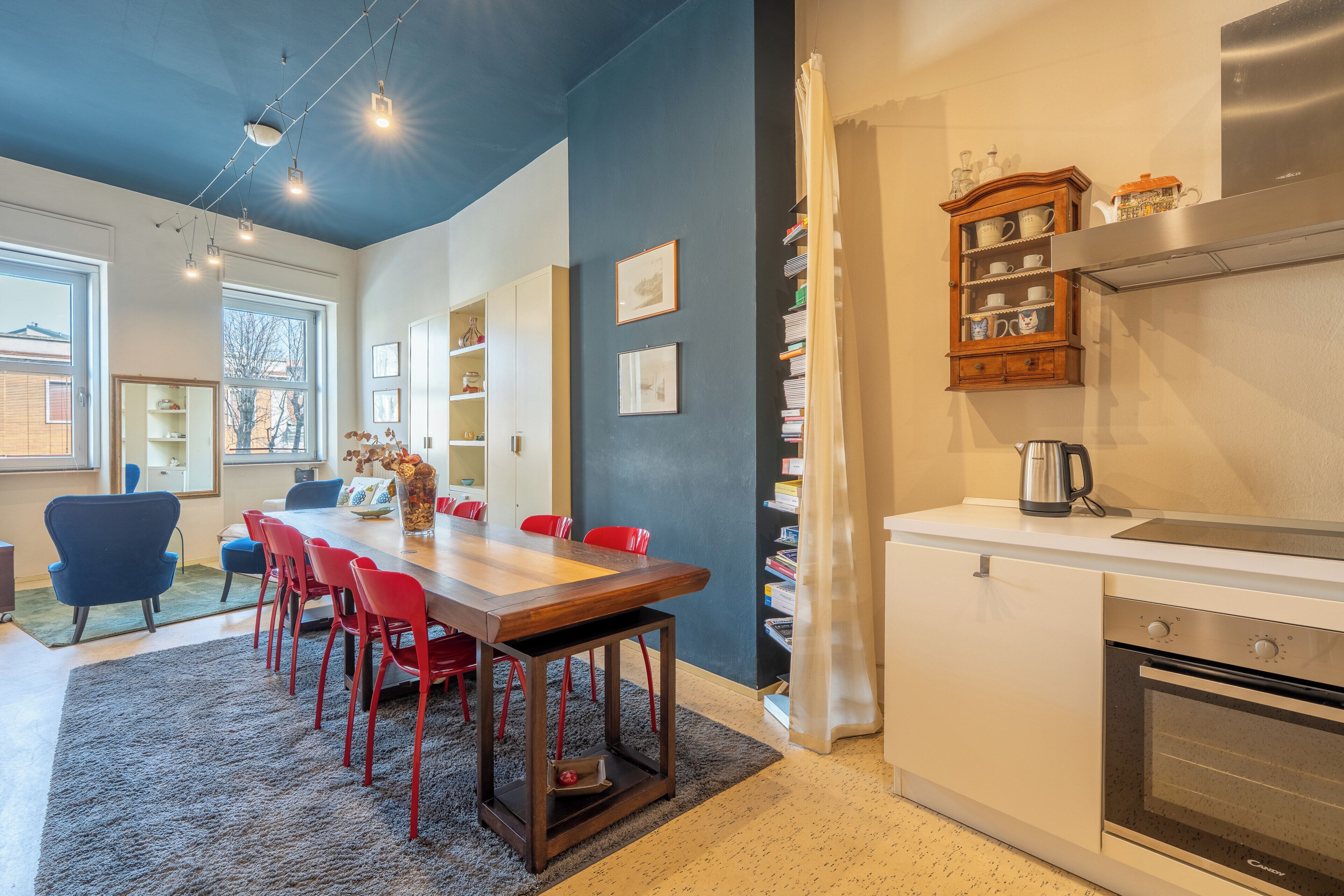 Una cucina moderna con forno a incasso, un'area pranzo con tavolo in legno e sedie rosse, e una libreria con diversi oggetti.