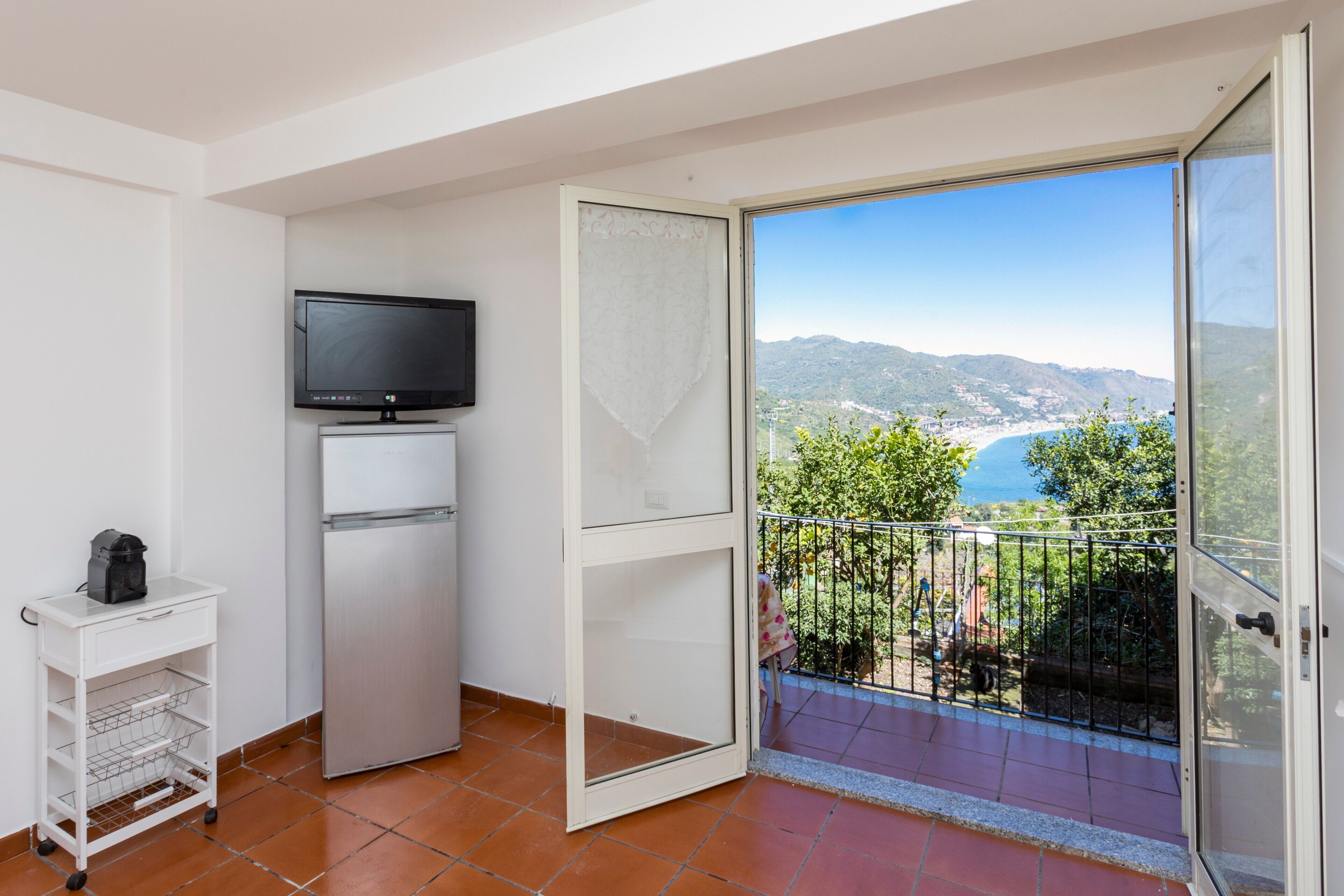A balcony with a clear view of the sea and mountains, a television on a white cabinet, and a white cart with a coffee maker.