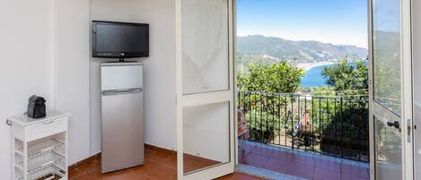 A balcony with a clear view of the sea and mountains, a television on a white cabinet, and a white cart with a coffee maker.