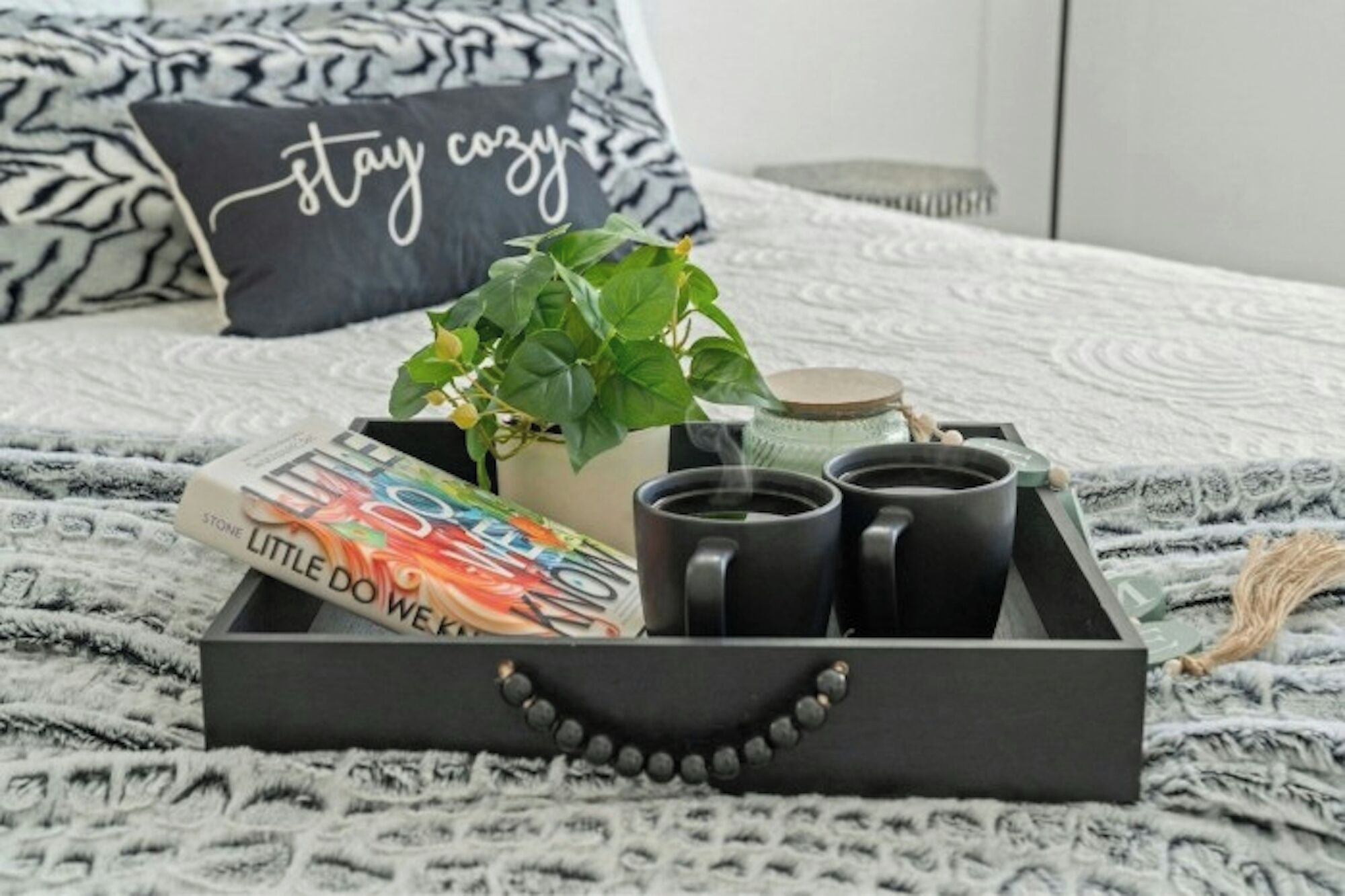 A bed with a black pillow, a potted plant, a book titled 'STONE LITTLE DOWEK,' and a tray with three black mugs.