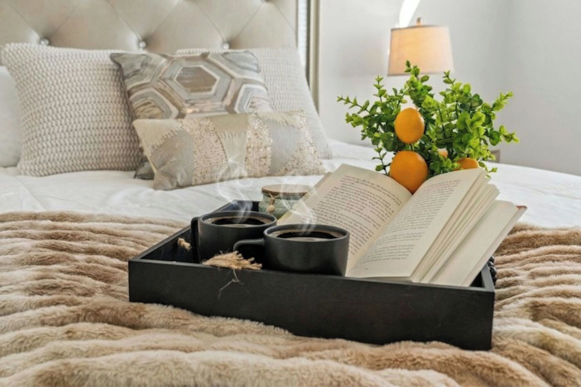 A bed with a black tray containing a book, two cups, and a small plant with orange fruits.