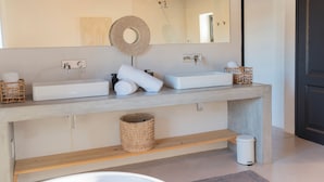 Un baño con un espejo grande, dos lavamanos, un estante de madera, un cubo de basura y un perchero para toallas.