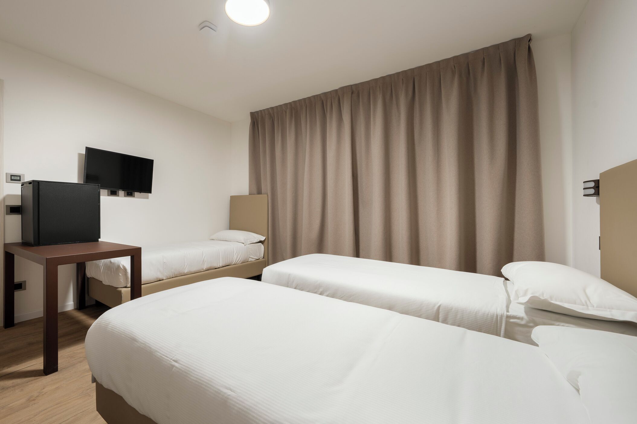 Ett hotellrum med två separata sängar, en TV, ett skrivbord och ett stort fönster med gardiner.