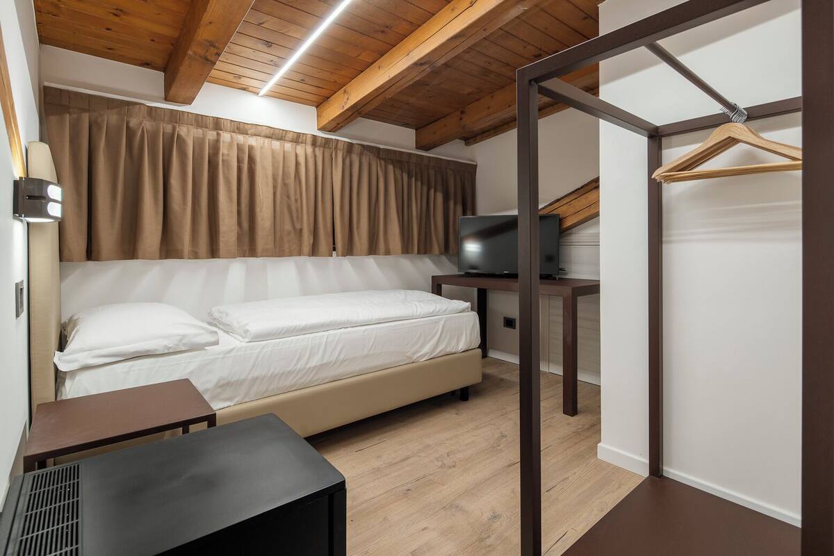 Ein Schlafzimmer mit einem Bett, einem Fernseher auf einem Standfuß, einer Holzbalkendecke und einem an der Wand befestigten Kleiderhaken.