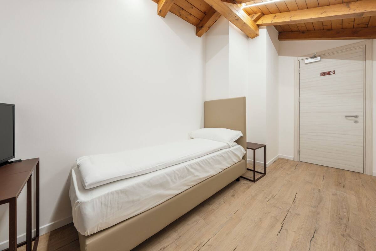Ein Einzelbett mit Kopfteil, ein kleiner Nachttisch, ein Fernseher auf einem Schrank und eine Tür mit einem Hinweisschild.
