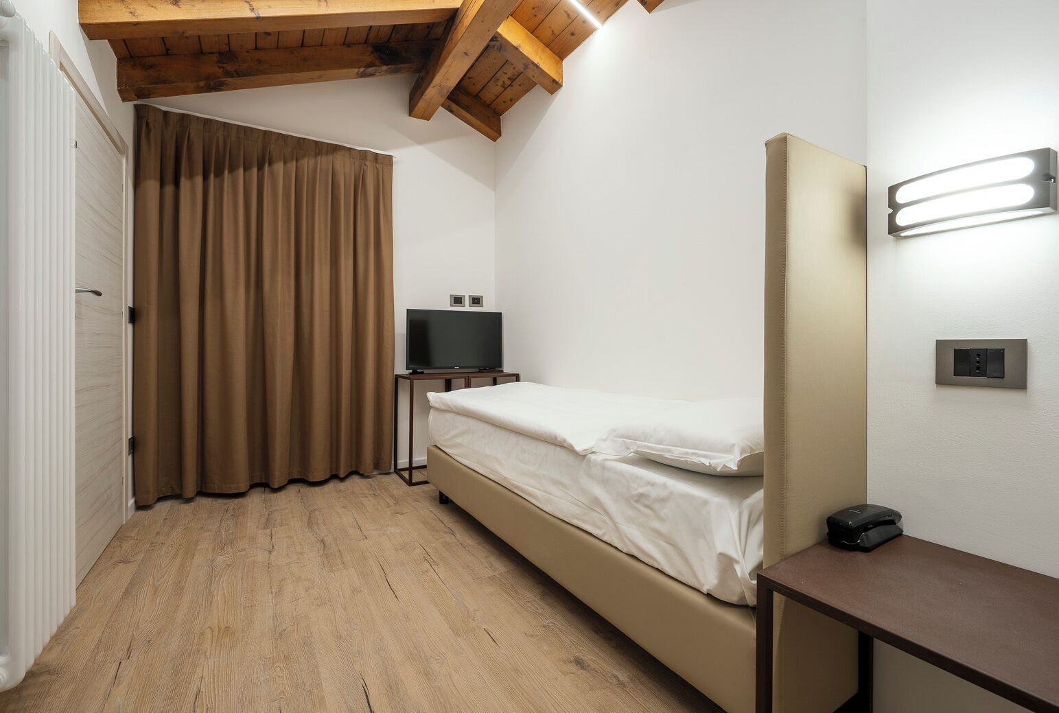 A small, single-bed room with a wooden ceiling, a television on a stand, a bedside table, and a wall-mounted light fixture.