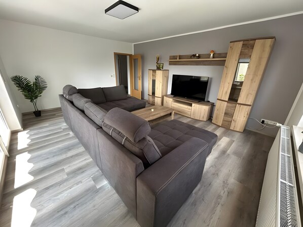 A modern living room with a sectional sofa, a flat-screen TV, wooden shelving, and a potted plant.