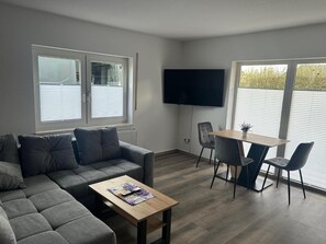 Ein modernes Wohnzimmer mit grauem Sofa, einem Holztisch, einem an der Wand befestigten Fernseher und einem Essbereich mit Tisch und Stühlen.