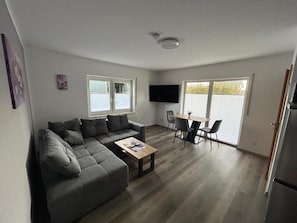 Un salon moderne avec un canapé d’angle gris, une table basse en bois, un téléviseur fixé au mur et un coin repas comprenant une table et des chaises.