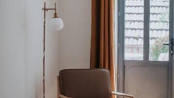 Una habitación con cama, una silla, una mesita, una lámpara de pie y un ventanal con cortinas.