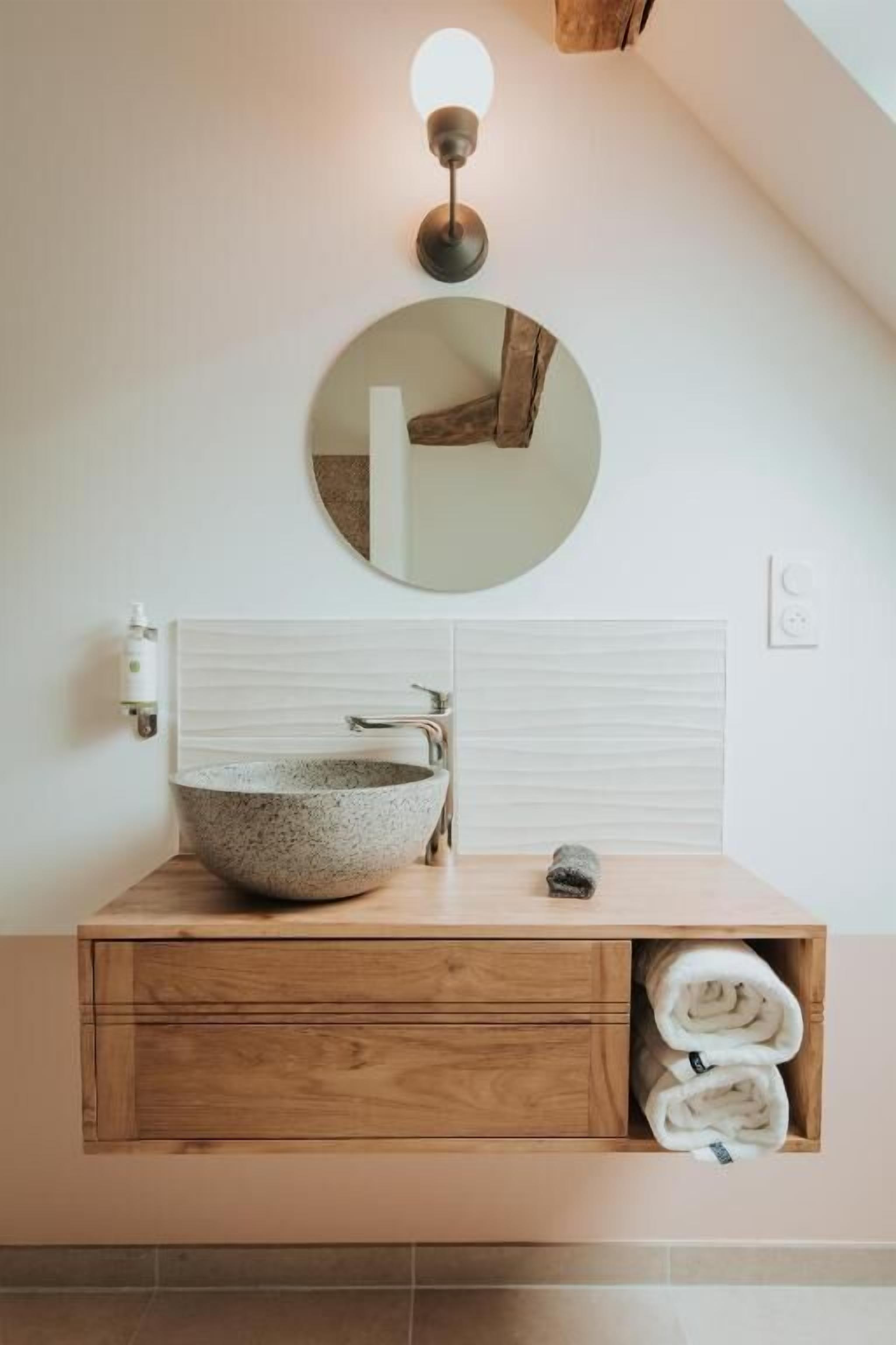 Un baño con un espejo redondo, un lavabo de piedra, tocador de madera y toallas.