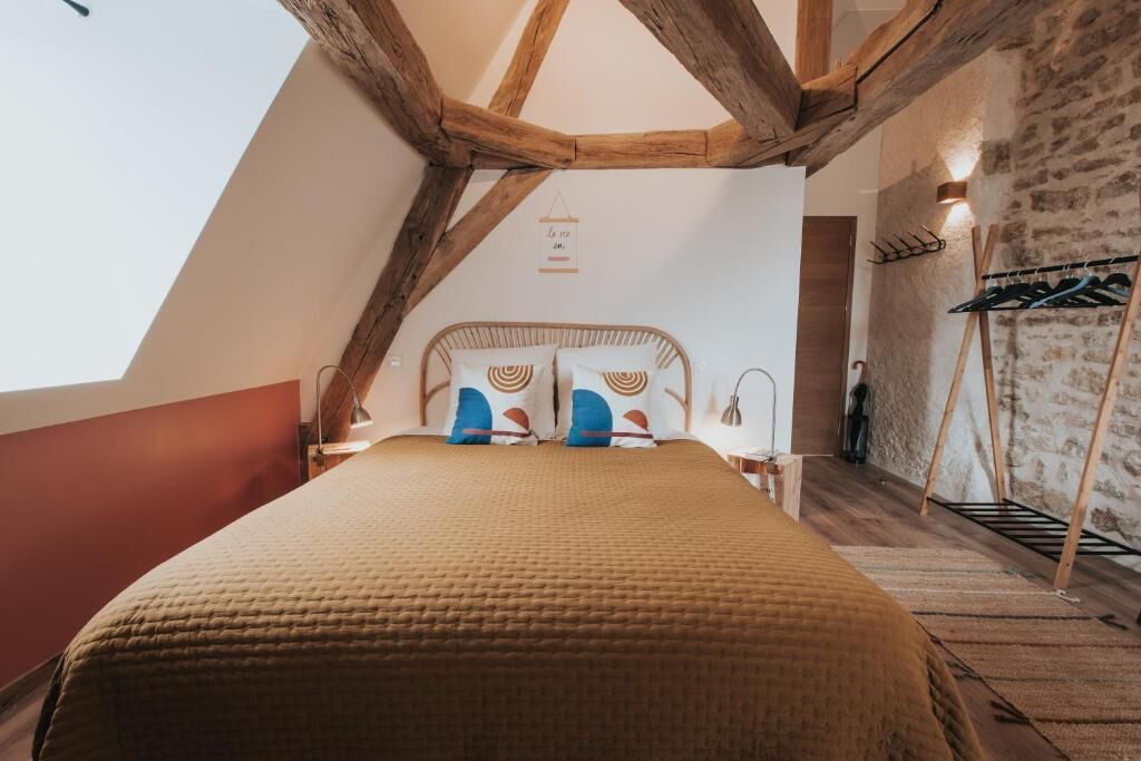 Ein Schlafzimmer mit schrägem Dach, einem Bett mit gemusterten Kissen, einer Wandleuchte und einem Holzregal mit Kleidung.