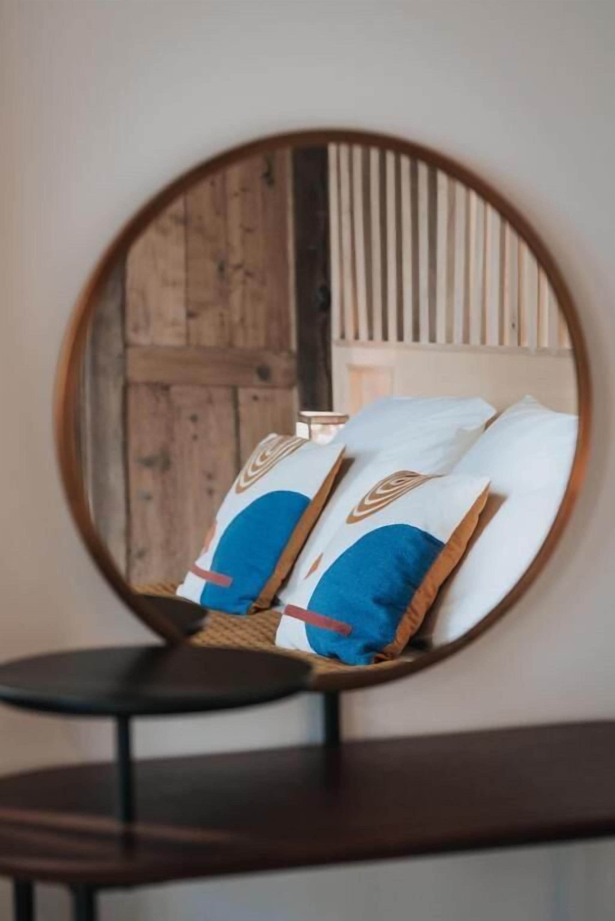 Un espejo redondo que refleja una cama con almohadas blancas y azules, una puerta de madera y una mesita pequeña.