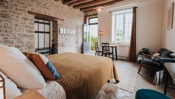 Ein Schlafzimmer mit Steinwand, Holzbalken, einem Bett mit Kissen, einem Schreibtisch mit Stuhl und einem Fenster mit Vorhängen.
