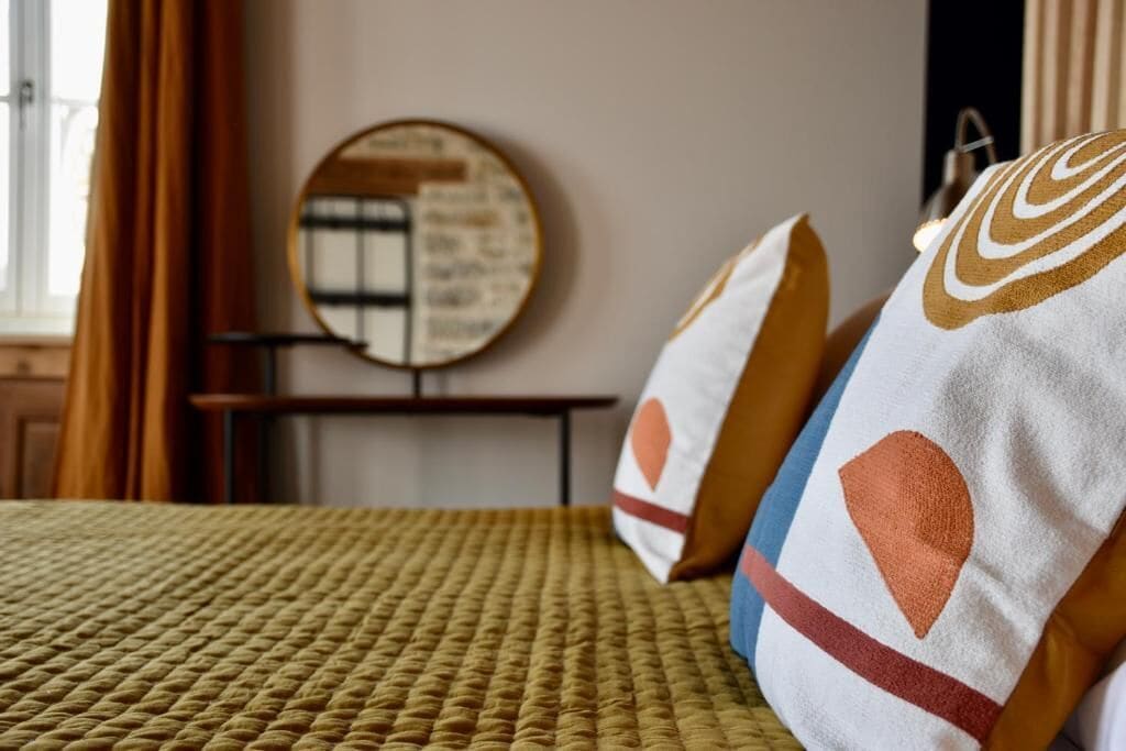 Un lit recouvert d’une couverture à motifs, des coussins aux motifs géométriques, un miroir rond accroché au mur et une fenêtre avec des rideaux.